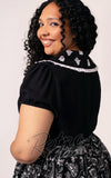 Woman wearing a black blouse with white lace details on a plain background curvy back